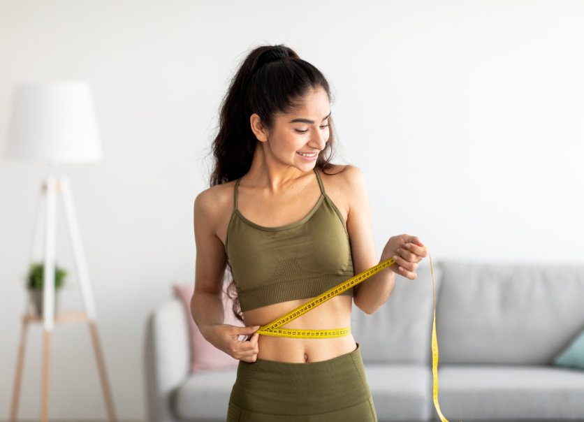 Fit young woman in sports clothes measuring waist with tape at home
